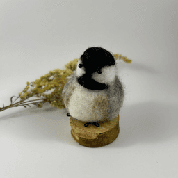 A small needle-felted black-capped chickadee sits on a half inch section of a branch, facing 1/4 towards the left. A dried spray of goldenrod lays in the background. Its face is white, its goatee black, its cap black, its breast white with tea-stain buff sides, its wings grey.