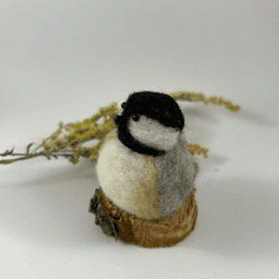 A small needle-felted black-capped chickadee sits on a half inch section of a branch, facing 3/4 towards the left. A dried spray of goldenrod lays in the background. Its face is white, its goatee black, its cap black, its breast white with tea-stain buff sides, its wings grey.