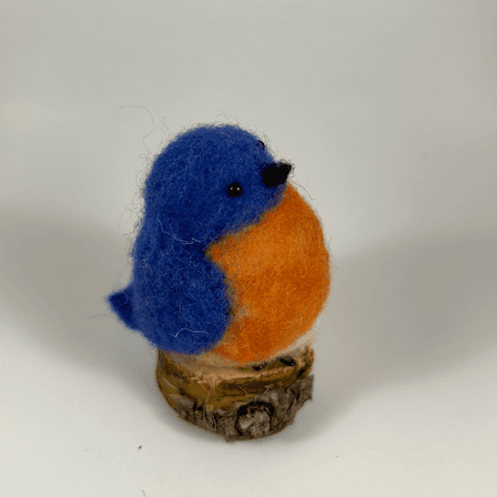 A needle-felted Eastern bluebird sits on a half inch section of a branch facing towards the right in profile. Its breast is bright orange with a white underside, its head, wings and tail royal blue.