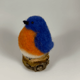 A needle-felted Eastern bluebird sits on a half inch section of a branch facing towards the left in 4/5 profile. Its breast is bright orange with a white underside, its head, wings and tail royal blue.