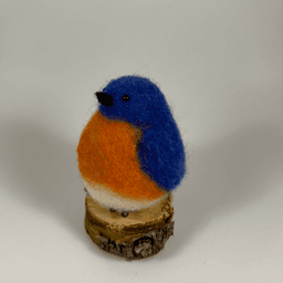 A needle-felted Eastern bluebird sits on a half inch section of a branch facing 3/4 towards the left. Its breast is bright orange with a white underside, its head, wings and tail royal blue.