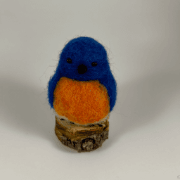 A needle-felted Eastern bluebird sits on a half inch section of a branch facing directly towards the camera. Its breast is bright orange with a white underside, its head, wings and tail royal blue.