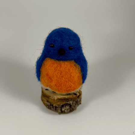 A needle-felted Eastern bluebird sits on a half inch section of a branch facing directly towards the camera. Its breast is bright orange with a white underside, its head, wings and tail royal blue.