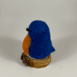 A needle-felted Eastern bluebird sits on a half inch section of a branch facing towards the left in total profile. Its breast is bright orange with a white underside, its head, wings and tail royal blue.