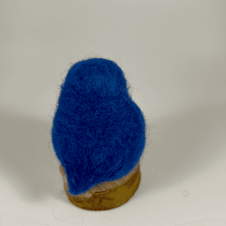 A needle-felted Eastern bluebird sits on a half inch section of a branch facing away from the camera. Its head, back, wings and tail are entirely royal blue.