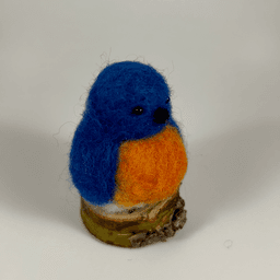 A needle-felted Eastern bluebird sits on a half inch section of a branch facing 1/4 towards the right. Its breast is bright orange with a white underside, its head, wings and tail royal blue.