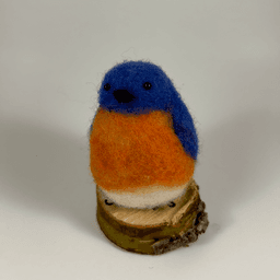 A needle-felted Eastern bluebird sits on a half inch section of a branch facing 1/4 towards the left. Its breast is bright orange with a white underside, its head, wings and tail royal blue.