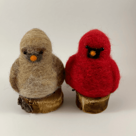 A pair of male and female Northern cardinals, scultped in needle felt, sit on half inch circle sections of branches, facing directly towards the camera. The female is a buff cream color with wine-stained wings, wine-stained eyebrows, a grey goatee and orange beak. The male is a cardinal red color with a black goatee and orange beak. Their crests are short and round.