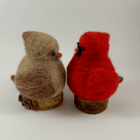 A pair of male and female Northern cardinals, scultped in needle felt, sit on half inch circle sections of branches, facing directly towards each other in profile. The female is a buff cream color with wine-stained wings, wine-stained eyebrows, a grey goatee and orange beak. The male is a cardinal red color with a black goatee and orange beak. Their crests and tail points are short and round.