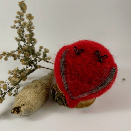 An upturned needle-felted male Cardinal sits revealing his flat red underside and grey outline underneath his wings, as well as black wire feets. There is a spray of goldenrod and a milkweed pod husk in the background.