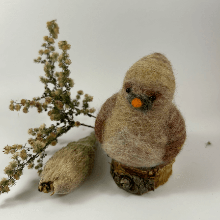 A female needle-felted Northern cardinal sits on a half-inch circle of cut branch, facing 1/4 towards the left. There is a spray of goldenrod and a milkweed pod husk in the background. Her body is a buff cream color, her wings and eyebrows are wine-stained, her goatee is grey, her beak orange. His crest is short and round.