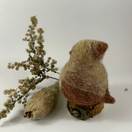 A female needle-felted Northern cardinal sits on a half-inch circle of cut branch, facing away from the camera 1/4 towards the left. There is a spray of goldenrod and a milkweed pod husk in the background. Her body is buff cream colored with wine-stained wings and tail, and wine-stained crest.