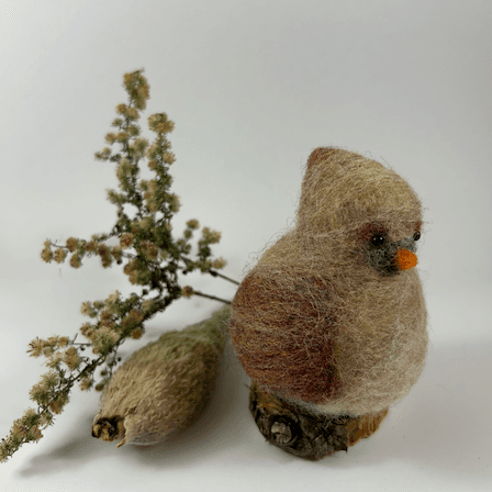 A female needle-felted Northern cardinal sits on a half-inch circle of cut branch, facing 3/4 towards the right. There is a spray of goldenrod and a milkweed pod husk in the background. Her body is a buff cream color, her wings, crest and tail are wine-stained, her beak orange. His crest is short and round.