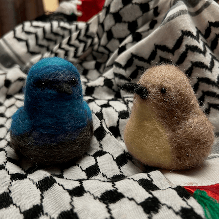 A pair of needle-felted Palestine sunbirds sit on the keffiyeh. The male faces directly towards the camera, his body ombre shades of bright and deep blues and dark, his proboscis beak black. The female faces 1/4 towards the left, her proboscis beak dark, her body buff colored and her belly a lighter buff color.