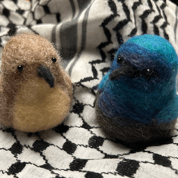 A pair of needle-felted Palestine sunbirds sit on the keffiyeh. The female on the left faces 1/4 towards the right, her proboscis beak dark, her body buff colored and her belly a lighter buff color. The male on the right faces 1/4 towards the left, his body ombre shades of bright and deep blues and dark, his proboscis beak black.