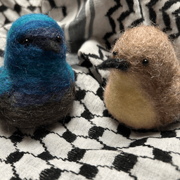 A pair of needle-felted Palestine sunbirds sit on the keffiyeh. The male on the left faces 1/4 towards the right, his body ombre shades of bright and deep blues and dark, his proboscis beak black.  The female on the right faces 1/4 towards the left, her proboscis beak dark, her body buff colored and her belly a lighter buff color.