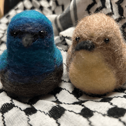 A pair of needle-felted Palestine sunbirds sit on the keffiyeh, facing almost directly towards the camera. The male on the left's body is ombre shades of bright and deep blues and dark, his proboscis beak black.  The female on the right has her proboscis beak dark, her body buff colored and her belly a lighter buff color.