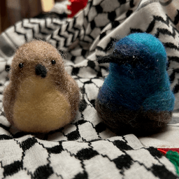 A pair of needle-felted Palestine sunbirds sit on the keffiyeh. The female on the left faces the camera directly, her proboscis beak dark, her body buff colored and her belly a lighter buff color.  The male on the right faces 1/4 towards the left, his body ombre shades of bright and deep blues and dark, his proboscis beak black.