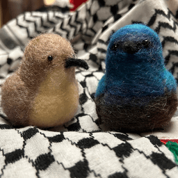 A pair of needle-felted Palestine sunbirds sit on the keffiyeh. The female on the left faces 1/4 towards the right, her proboscis beak dark, her body buff colored and her belly a lighter buff color.  The male on the right faces directly towards the camera, his body ombre shades of bright and deep blues and dark, his proboscis beak black.