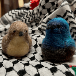 A pair of needle-felted Palestine sunbirds sit on the keffiyeh. The female on the left faces directly towards the camera, her proboscis beak dark, her body buff colored and her belly a lighter buff color.  The male on the right faces 1/4 towards the left, his body ombre shades of bright and deep blues and dark, his proboscis beak black.