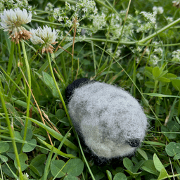 A needle-felted horned sheep with black head, horns and legs and light grey fleece faces away from the camera 1/4 towards the left. The sheep sits on clover, with flowering clovers in the background. Its small round black tail emerges from its wooly behind.