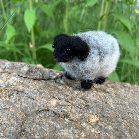 A needle-felted horned sheep with black head, horns and legs and light grey fleece faces 3/4 towards the left. The sheep stands on a large rock with green growing in the background.