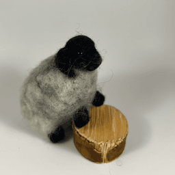 A needle-felted horned sheep with black head, horns and legs and light grey fleece stands its forelegs on a half inch thick cut section of branch, facing 3/4 towards the right.