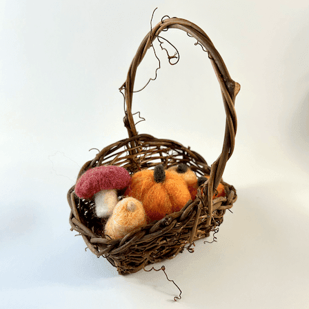 A small viney basket holds needle felted produce: a mushroom, a butternut squash, and three pumpkins of different shades of orange.