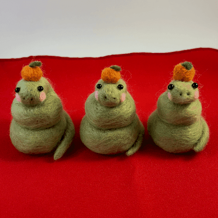 Three green needle-felted snakes curled up with pink cheeks wearing a mandarin orange on their head. They are seated on a red cloth and facing the camera.