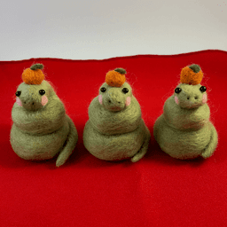 Three green needle-felted snakes curled up with pink cheeks wearing a mandarin orange on their head. They are seated on a red cloth and facing the camera.