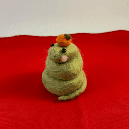 A green needle-felted snake with pink cheeks curled up wearing a mandarin orange on its head faces 1/4 to the left. It sits on a red cloth.