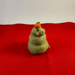 A green needle-felted snake with pink cheeks curled up wearing a mandarin orange on its head faces 1/4 to the right. It sits on a red cloth.