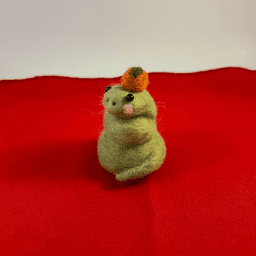 A green needle-felted snake with pink cheeks curled up wearing a mandarin orange on its head faces 1/4 to the left. It sits on a red cloth.