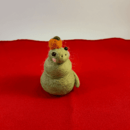 A green needle-felted snake with pink cheeks curled up wearing a mandarin orange on its head faces slightly to the right. It sits on a red cloth.