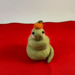 A green needle-felted snake with pink cheeks curled up wearing a mandarin orange on its head faces directly towards the camera. It sits on a red cloth.