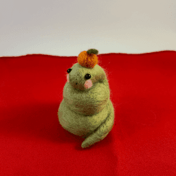 A green needle-felted snake with pink cheeks curled up wearing a mandarin orange on its head faces slightly to the left. It sits on a red cloth.