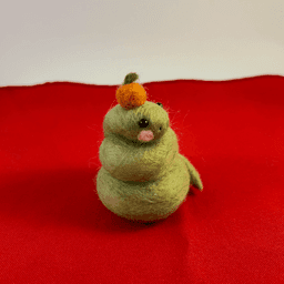 A green needle-felted snake with pink cheeks curled up wearing a mandarin orange on its head faces 1/4 to the right. It sits on a red cloth.