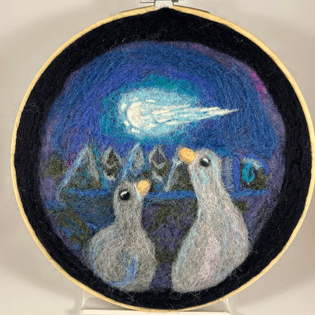 Wool painting of two ducks watching a meteor in the night sky, the light reflected in their awe-filled eyes. The painting is cool-toned with mostly dark blues, and details in purple, pink, and light blue. The wool painting sits in an 8 inch embroidery frame.