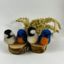 Four miniature needle-felted birds sit on two half-inch cross sections of a branch, sitting in alternating species: Eastern blue bird and black-capped chickadee. Two birds fit on each branch cut out. There is a spray of goldenrod in the background.