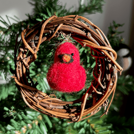 A small four inch diameter vine wreath with a needle-felted tiny Northern Cardinal hanging in its center adorns a small artificial Christmas tree.