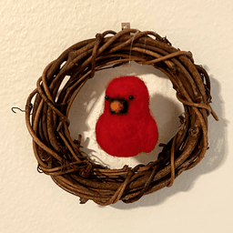 A small four inch diameter vine wreath with a needle-felted tiny Northern Cardinal hanging in its center hands on an off white colored wall.