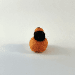 A tiny needle-felted black corvid borb pokes its head out of an orange pumpkin, wearing a pumpkin stem hat. The mini borb faces directly towards the camera.