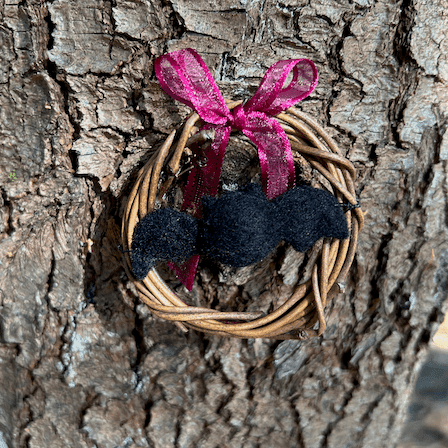 Miniature wreath hangs on a pine tree. In its center hangs a needle felted black bat. A wine colored translucent bow is tied at the top of the wreath.