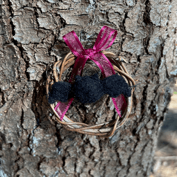 Miniature wreath hangs on a pine tree. In its center hangs a needle felted black bat. A wine colored translucent bow is tied at the top of the wreath.