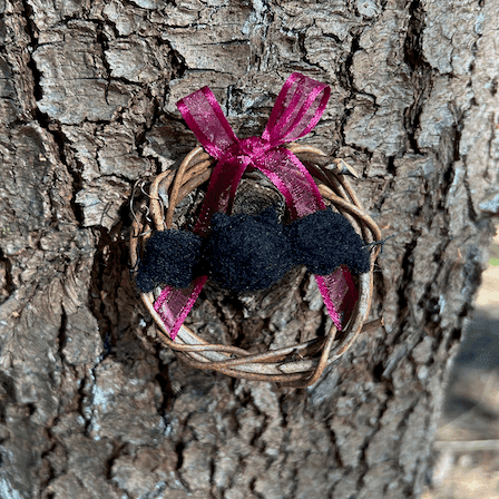 Miniature wreath hangs on a pine tree. In its center hangs a needle felted black bat. A wine colored translucent bow is tied at the top of the wreath.