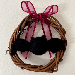 Miniature wreath hangs on a wall. In its center hangs a needle felted black bat. A wine colored translucent bow is tied at the top of the wreath.