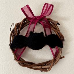 Miniature wreath hangs on a wall. In its center hangs a needle felted black bat. A wine colored translucent bow is tied at the top of the wreath.