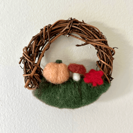 Miniature wreath hangs on a white wall. On its bottom is green felting wool topped with a light orange pumpkin, a mushroom, and a red maple leaf.