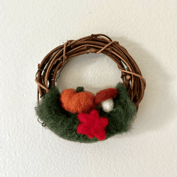 Miniature wreath hangs on a white wall. On its bottom is green felting wool topped with an orange pumpkin, a mushroom, and a red maple leaf in center.