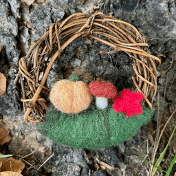 Miniature wreath sits at the base of a birch tree. On its bottom is green felting wool topped with a light orange pumpkin, a mushroom, and a red maple leaf.